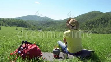 那个拿着旅游棒和红色背包的旅行者女人坐在草地上，煤气炉上的茶壶站着